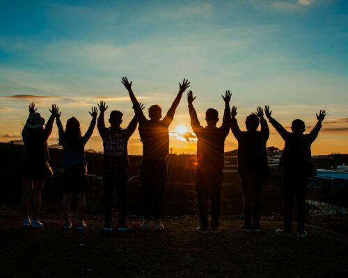 A diverse group of friends raises their arms in celebration against a vibrant sunset backdrop.
